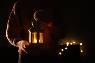 Photo of Woman holding Christmas lantern with burning candle in darkness, closeup. Bokeh effect