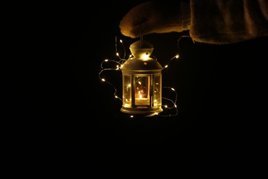 Photo of Woman holding Christmas lantern with burning candle and fairy lights in darkness, closeup. Space for text