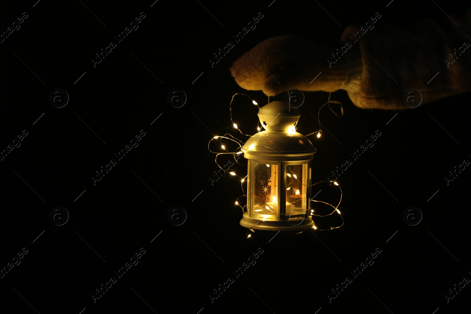 Photo of Woman holding Christmas lantern with burning candle and fairy lights in darkness, closeup. Space for text