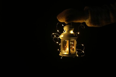 Photo of Woman holding Christmas lantern with burning candle and fairy lights in darkness, closeup. Space for text