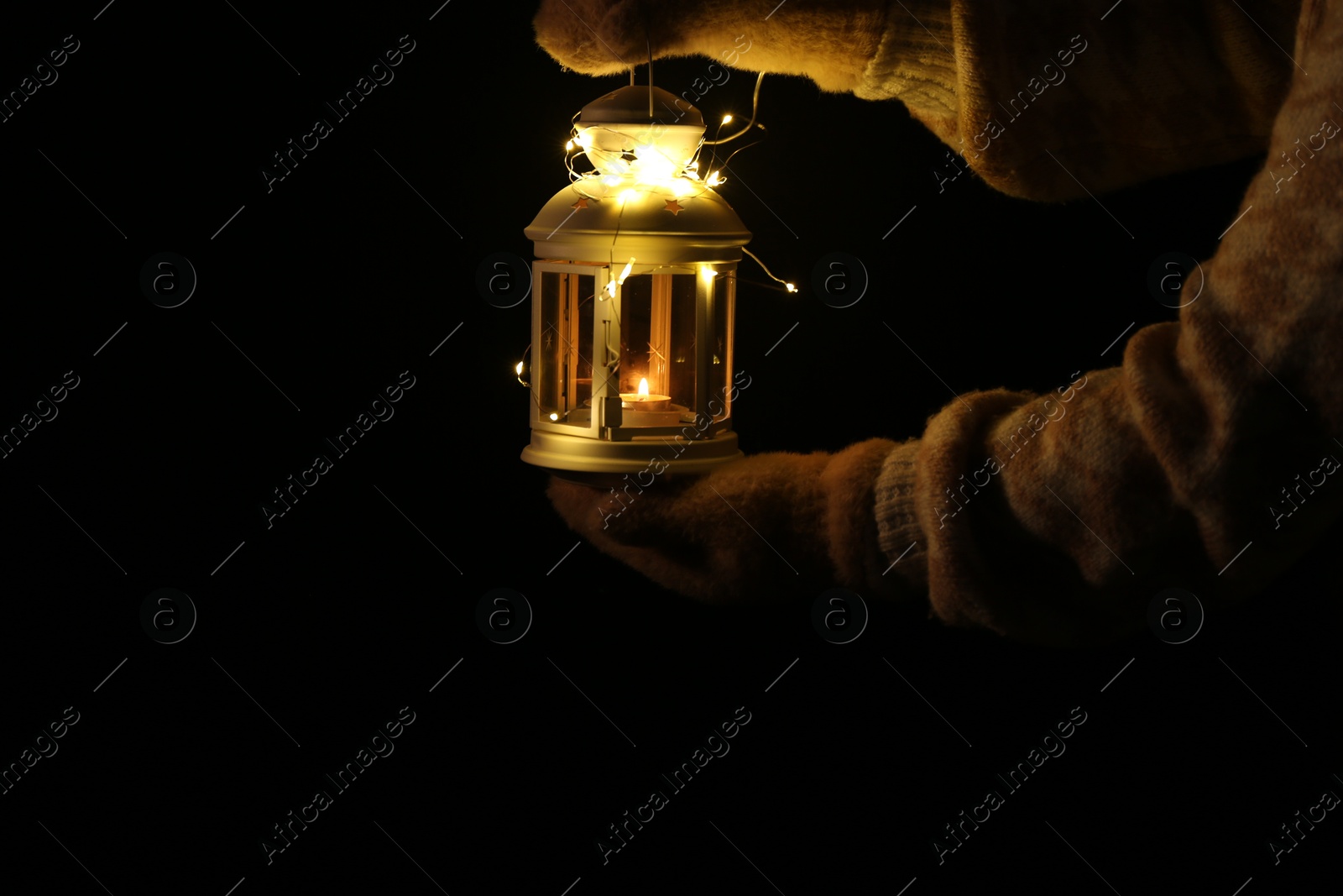 Photo of Woman holding Christmas lantern with burning candle and fairy lights in darkness, closeup. Space for text