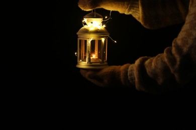 Photo of Woman holding Christmas lantern with burning candle and fairy lights in darkness, closeup. Space for text