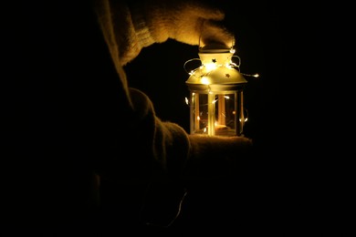 Photo of Woman holding Christmas lantern with burning candle and fairy lights in darkness, closeup. Space for text