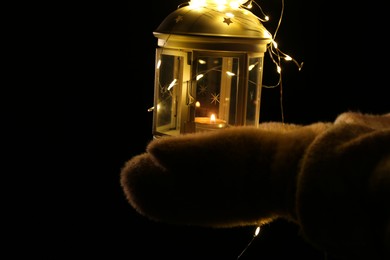 Photo of Woman holding Christmas lantern with burning candle and fairy lights in darkness, closeup. Space for text