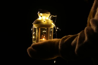 Photo of Woman holding Christmas lantern with burning candle and fairy lights in darkness, closeup