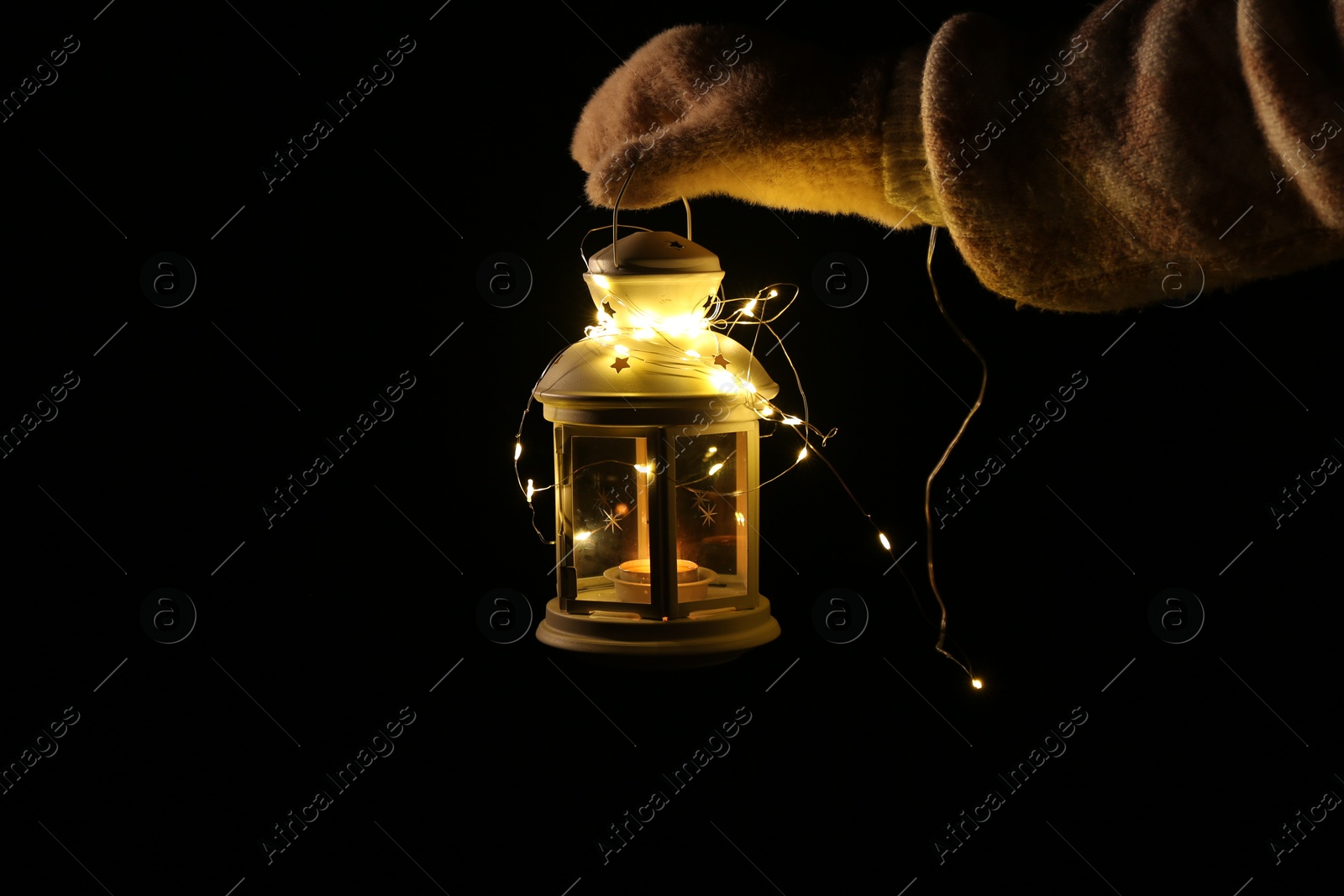 Photo of Woman holding Christmas lantern with burning candle and fairy lights in darkness, closeup. Space for text