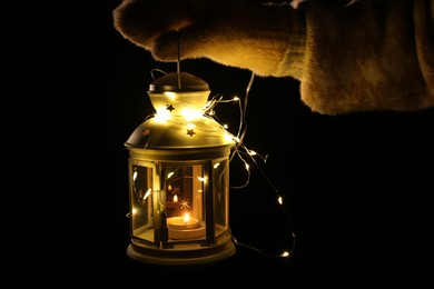 Photo of Woman holding Christmas lantern with burning candle and fairy lights in darkness, closeup