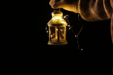 Photo of Woman holding Christmas lantern with burning candle and fairy lights in darkness, closeup. Space for text