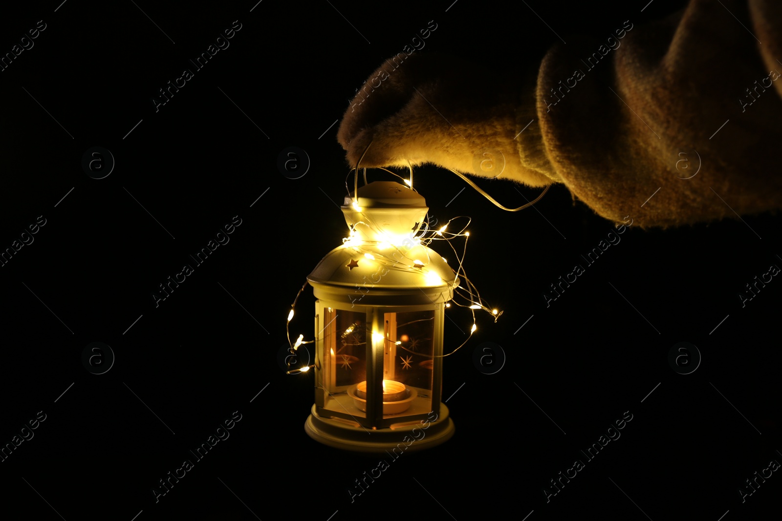 Photo of Woman holding Christmas lantern with burning candle and fairy lights in darkness, closeup. Space for text