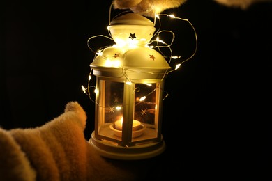 Photo of Woman holding Christmas lantern with burning candle and fairy lights in darkness, closeup