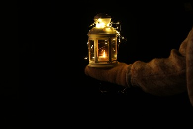 Photo of Woman holding Christmas lantern with burning candle and fairy lights in darkness, closeup. Space for text