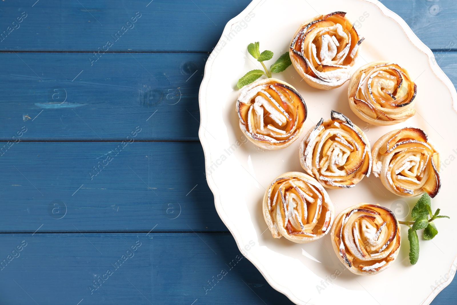 Photo of Tasty apple roses with powdered sugar served on blue wooden table, top view