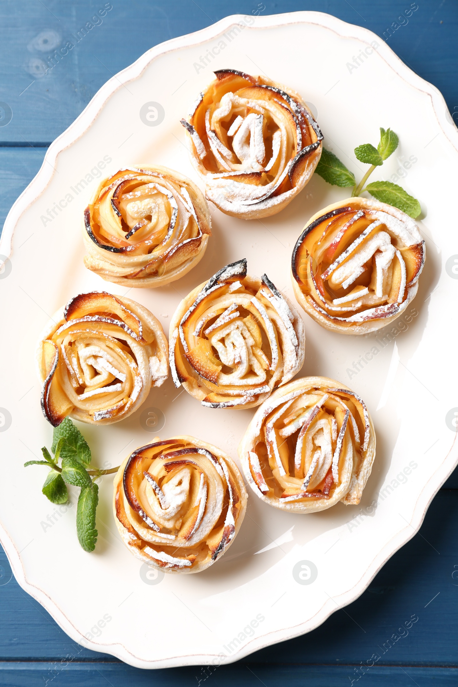 Photo of Tasty apple roses with powdered sugar served on blue wooden table, top view