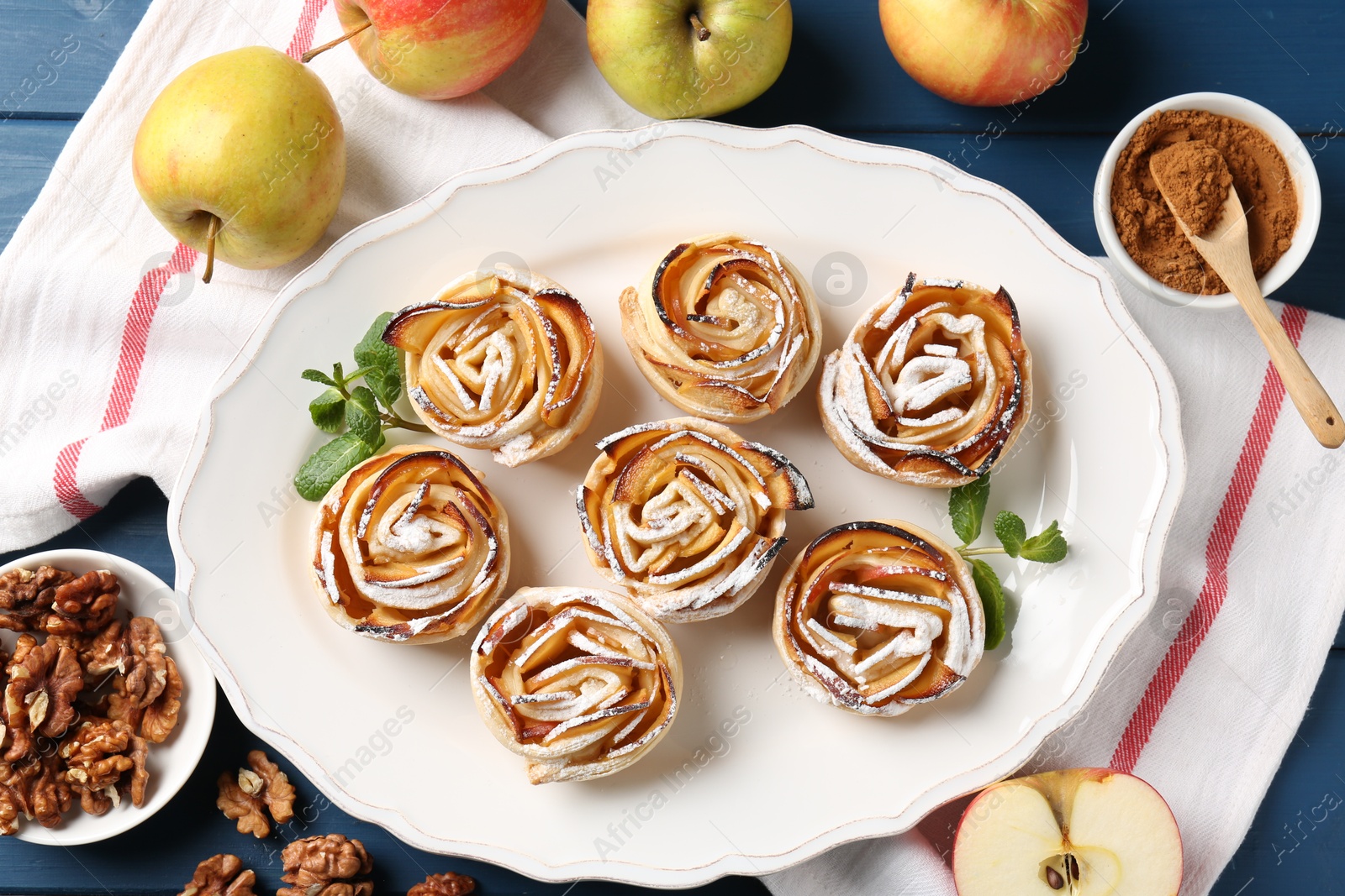 Photo of Tasty apple roses with powdered sugar served on blue wooden table, flat lay