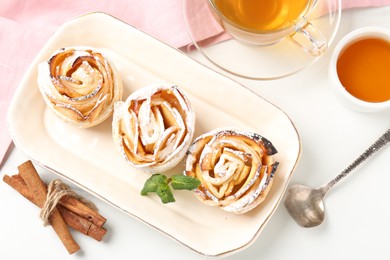 Photo of Freshly baked apple roses with mint, honey, cinnamon sticks and tea on white table, flat lay