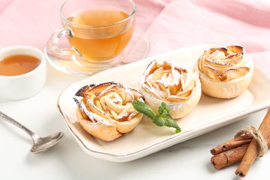Photo of Freshly baked apple roses with mint, honey, cinnamon sticks and tea on white marble table, closeup