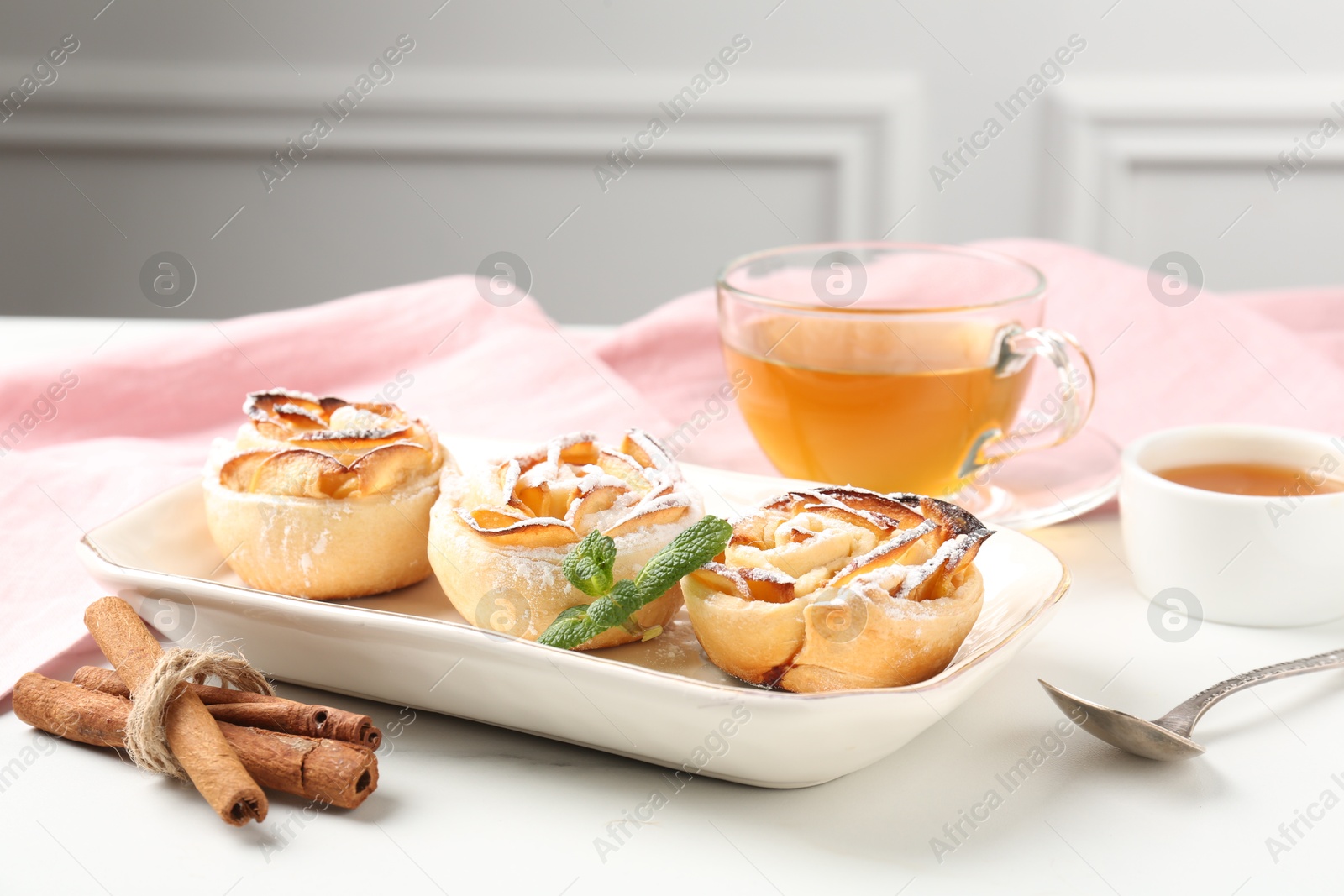 Photo of Freshly baked apple roses with mint, honey, cinnamon sticks and tea on white marble table, closeup