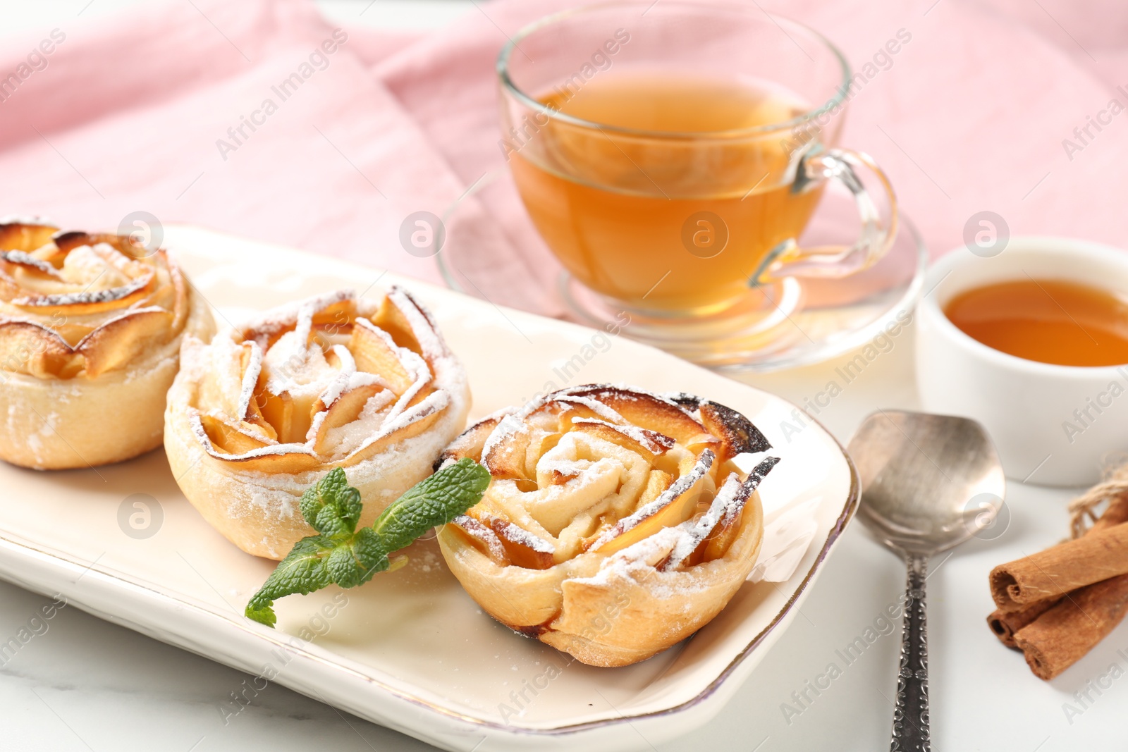 Photo of Freshly baked apple roses with mint, honey, cinnamon sticks and tea on white marble table, closeup