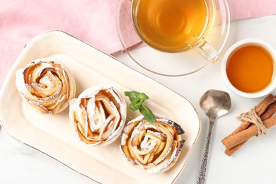 Photo of Freshly baked apple roses with mint, honey, cinnamon sticks and tea on white marble table, flat lay