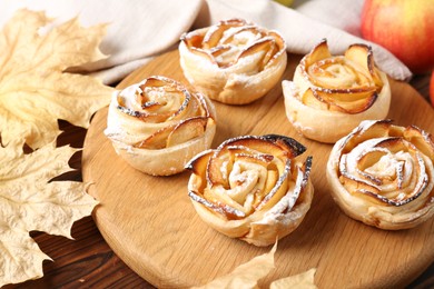 Photo of Freshly baked apple roses and dry leaves on wooden table, closeup