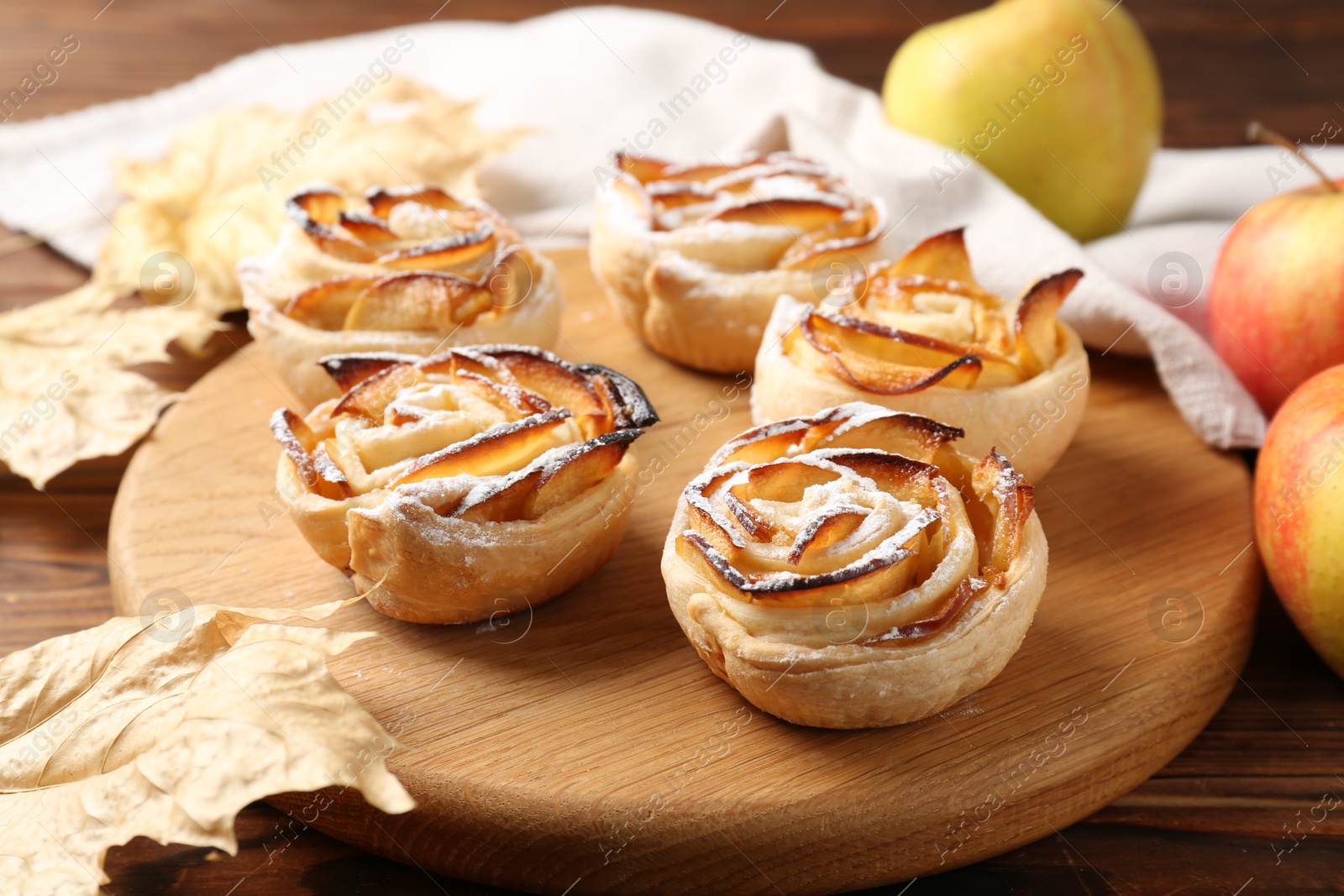 Photo of Freshly baked apple roses, fruits and dry leaves on wooden table, closeup