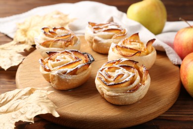 Photo of Freshly baked apple roses, fruits and dry leaves on wooden table, closeup