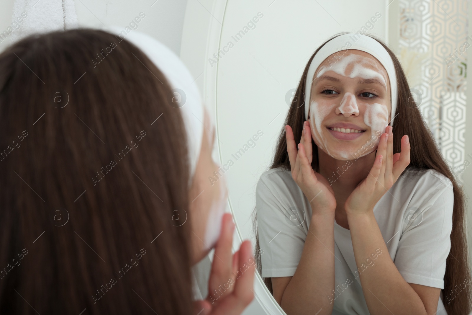 Photo of Teenage girl applying cleansing foam onto her face near mirror indoors. Acne treatment