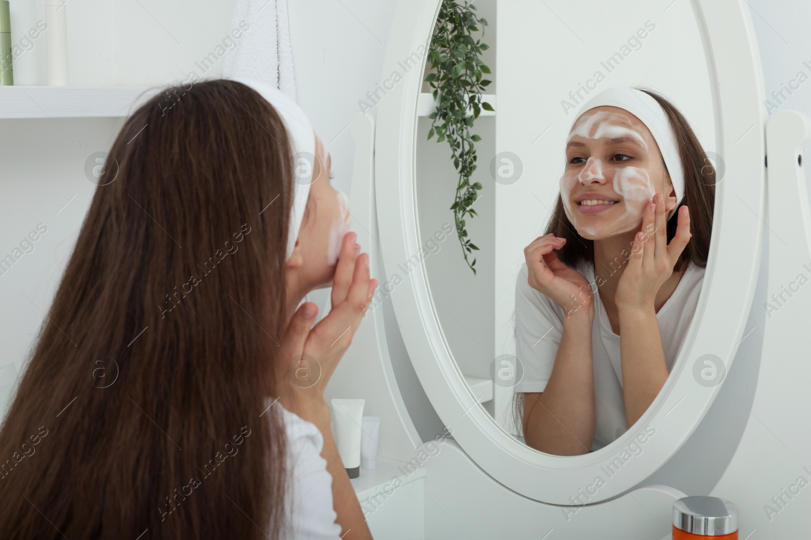 Photo of Teenage girl applying cleansing foam onto her face near mirror indoors. Acne treatment