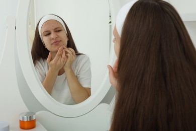 Photo of Upset teenage girl popping pimple on her face near mirror indoors. Acne problem