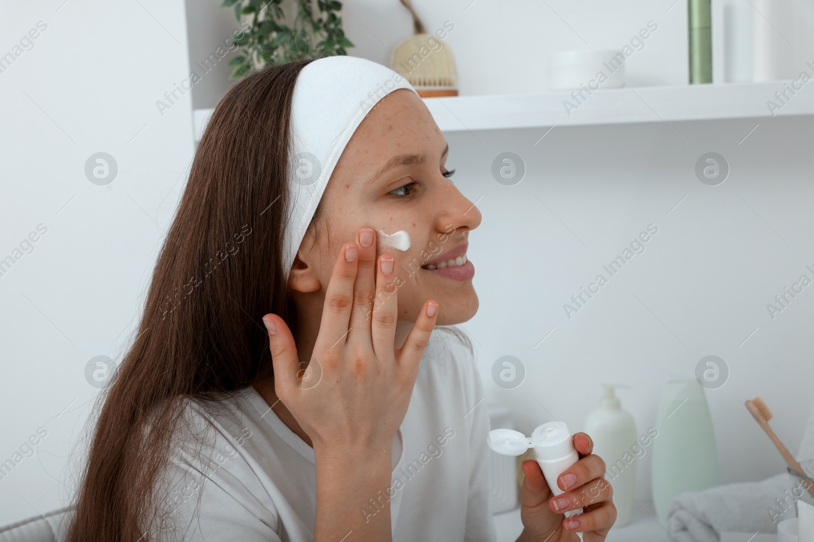 Photo of Teenage girl applying cream onto face near mirror indoors. Acne treatment