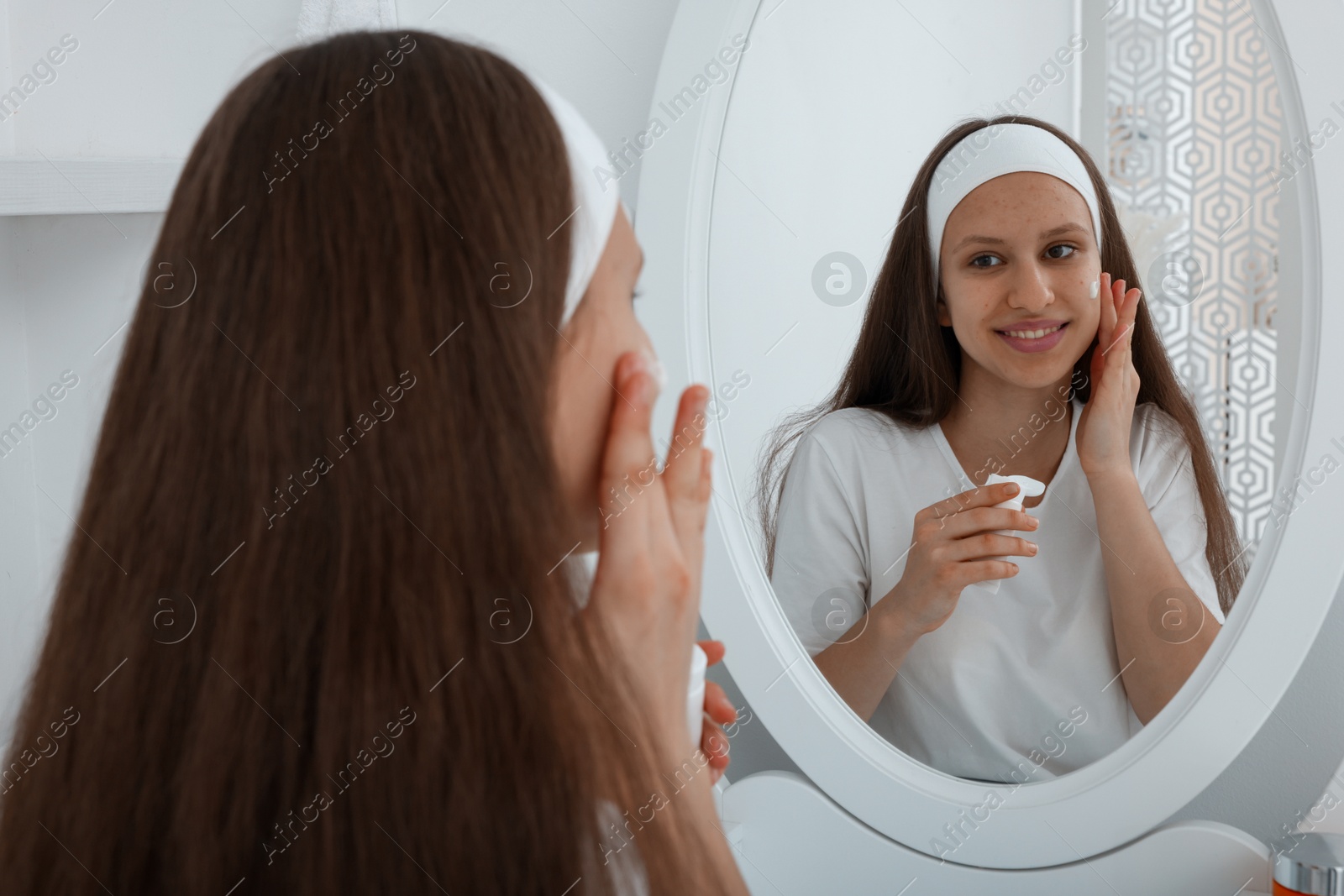 Photo of Teenage girl applying cream onto face near mirror indoors. Acne treatment