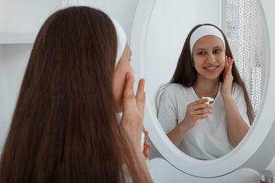 Photo of Teenage girl applying cream onto face near mirror indoors. Acne treatment