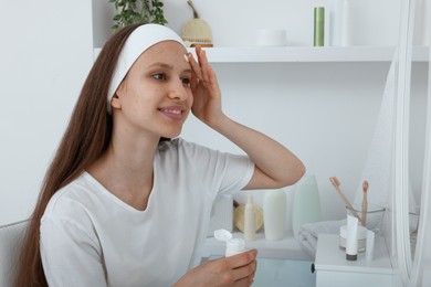 Photo of Teenage girl applying cream onto face near mirror indoors. Acne treatment