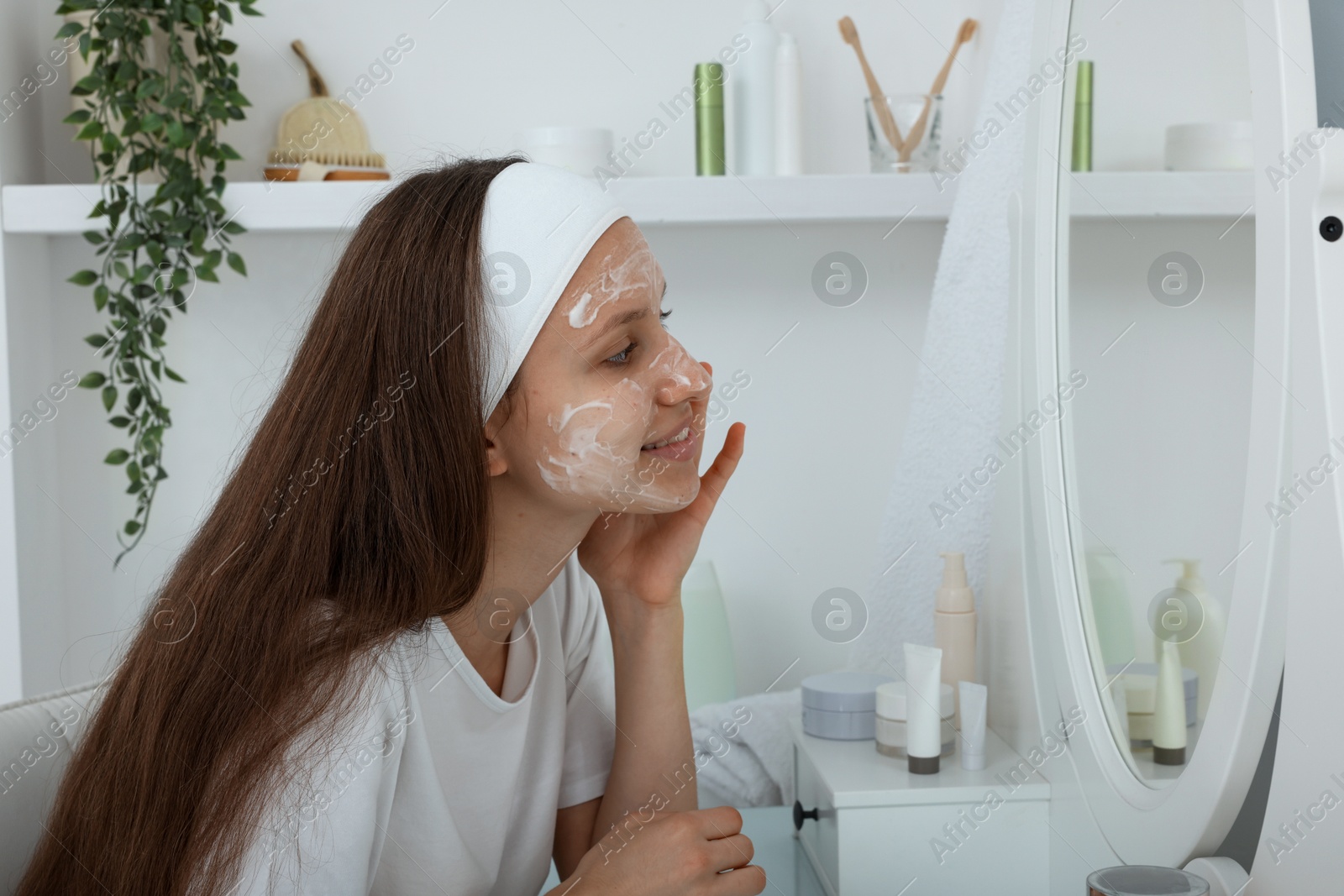 Photo of Teenage girl applying cream onto face near mirror indoors. Acne treatment