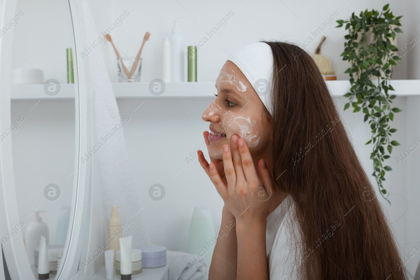 Photo of Teenage girl applying cream onto face near mirror indoors. Acne treatment