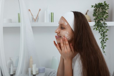 Photo of Teenage girl applying cream onto face near mirror indoors. Acne treatment