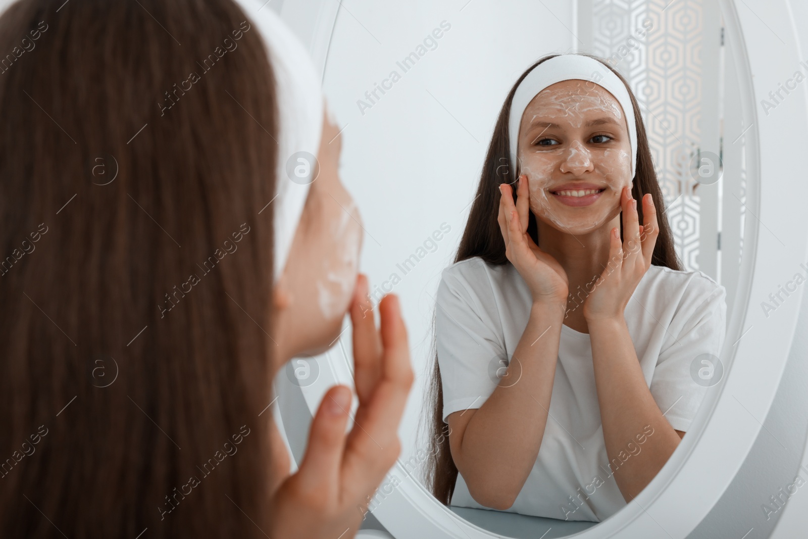 Photo of Teenage girl applying cream onto face near mirror indoors. Acne treatment