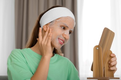 Photo of Teenage girl with sheet facial mask looking in mirror indoors. Acne treatment