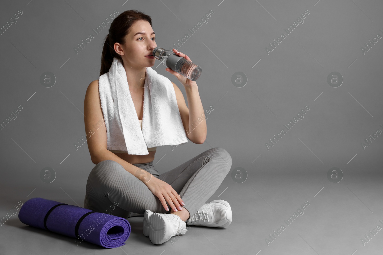 Photo of Woman in sportswear with towel and fitness mat drinking water on grey background, space for text