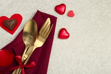 Romantic place setting for Valentine's day. Cutlery, napkin, candy, petals and decorative hearts on light textured table, flat lay. Space for text