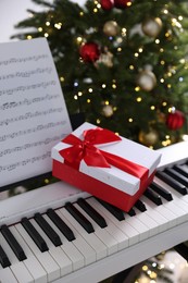 Synthesizer with music sheet and gift box near Christmas tree indoors, closeup