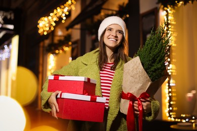 Happy woman with thuja tree and Christmas gifts outdoors, bokeh effect