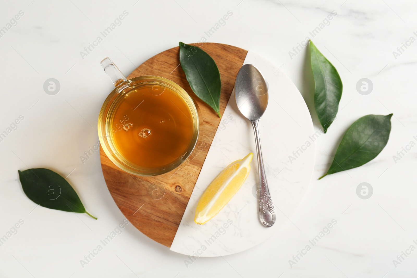 Photo of Refreshing green tea in cup, spoon, leaves and slice of lemon on white marble table, flat lay