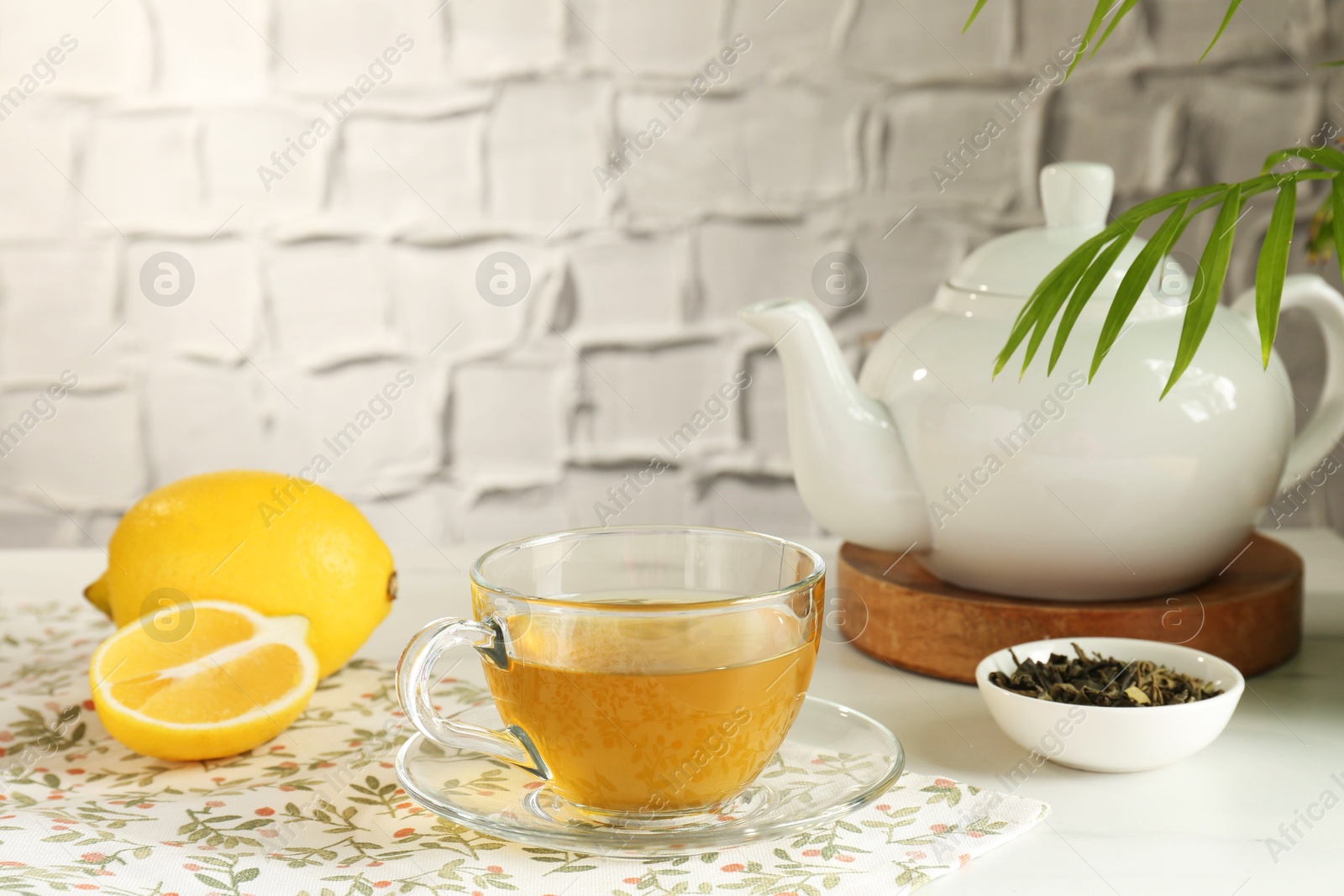 Photo of Refreshing green tea in cup, lemon and dry leaves on white table against textured wall