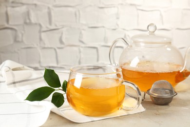 Photo of Refreshing green tea, strainer and leaves on light table against textured wall, closeup