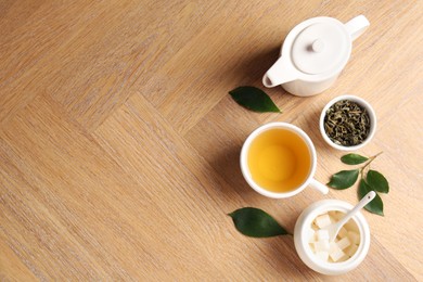 Photo of Refreshing green tea in cup, teapot, sugar bowl and leaves on wooden table, flat lay. Space for text