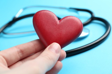 Photo of Cardiology. Woman with red decorative heart and stethoscope on light blue background, closeup