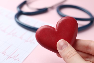 Photo of Cardiology. Woman with red decorative heart, cardiogram and stethoscope on pale pink background, closeup