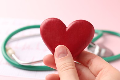 Photo of Cardiology. Woman with red decorative heart, cardiogram and stethoscope on pale pink background, closeup