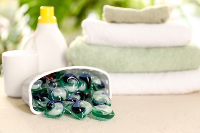 Photo of Container with laundry capsules, detergents and clean towels on table against blurred background, selective focus